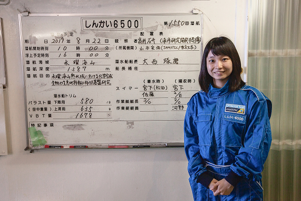 写真：青い作業服を着た山本実侑氏が、潜水艇「しんかい6500」の潜航内容が書かれた掲示板の前に立っている様子