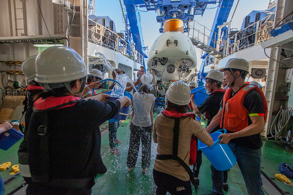 写真：潜水艇「しんかい6500」が引き上げられる様子。乗組員に水をかけるクルーたち。白いヘルメットと救命胴衣を着用