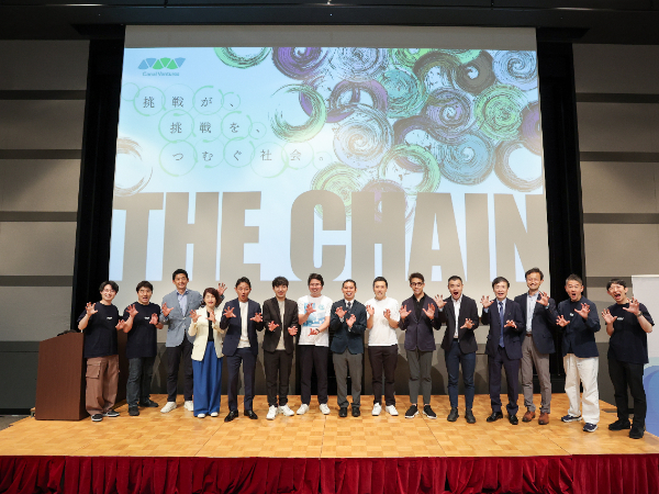 写真：「挑戦が、挑戦をつむぐ社会。THE CHAIN」のスクリーンを背景に、関係者一同がステージ上で並んで記念撮影をしている様子
