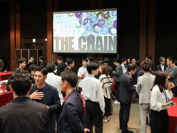 写真：「THE CHAIN」のロゴが表示された大型スクリーンの前で、多数の参加者が交流会で歓談している様子