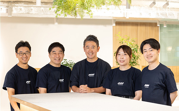 写真：ロゴ入りTシャツを着た男性4名と女性1名のスタッフ5名が、テーブルを囲んで笑顔で並んで写っている様子