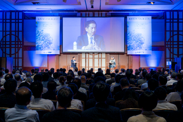 写真：BIPROGY FORUM 2026の会場で、大型スクリーンに登壇者の様子が映し出され、多くの参加者が着席して対談の様子を見つめている様子