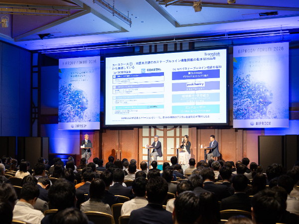写真：講演会場の様子。壇上のスクリーンにはスライドが表示され、4名の登壇者が並んで語り合っており、多くの参加者が着席して聴講している。