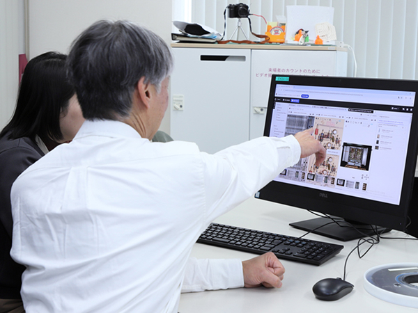 写真：デスクトップPCの画面を見ながら、男性が指で画像を示して説明している様子。隣に別の人物が座って一緒に確認している。