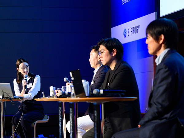 写真：パネルディスカッションの様子。司会者がマイクを手に登壇者へ問いかける。背後のスクリーンには「BIPROGY」のロゴが表示されている。